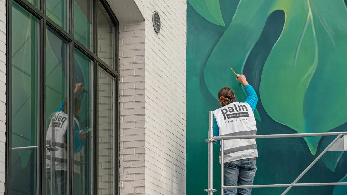 A muralist on scaffolding hand-painting a large green botanical mural